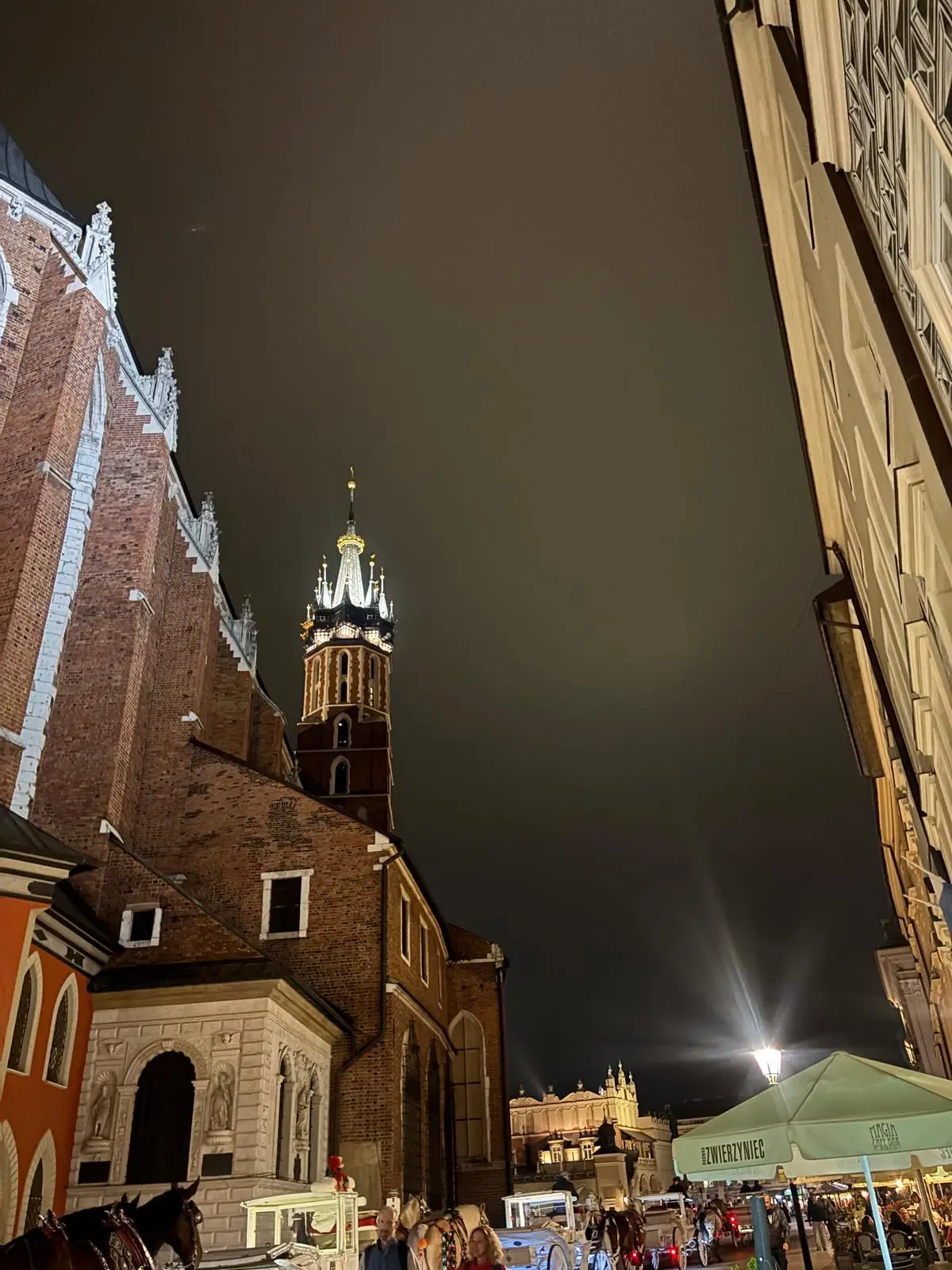 La torre de la basílica de Santa María de Cracovia iluminada de noche, con coches de caballos en la plaza
